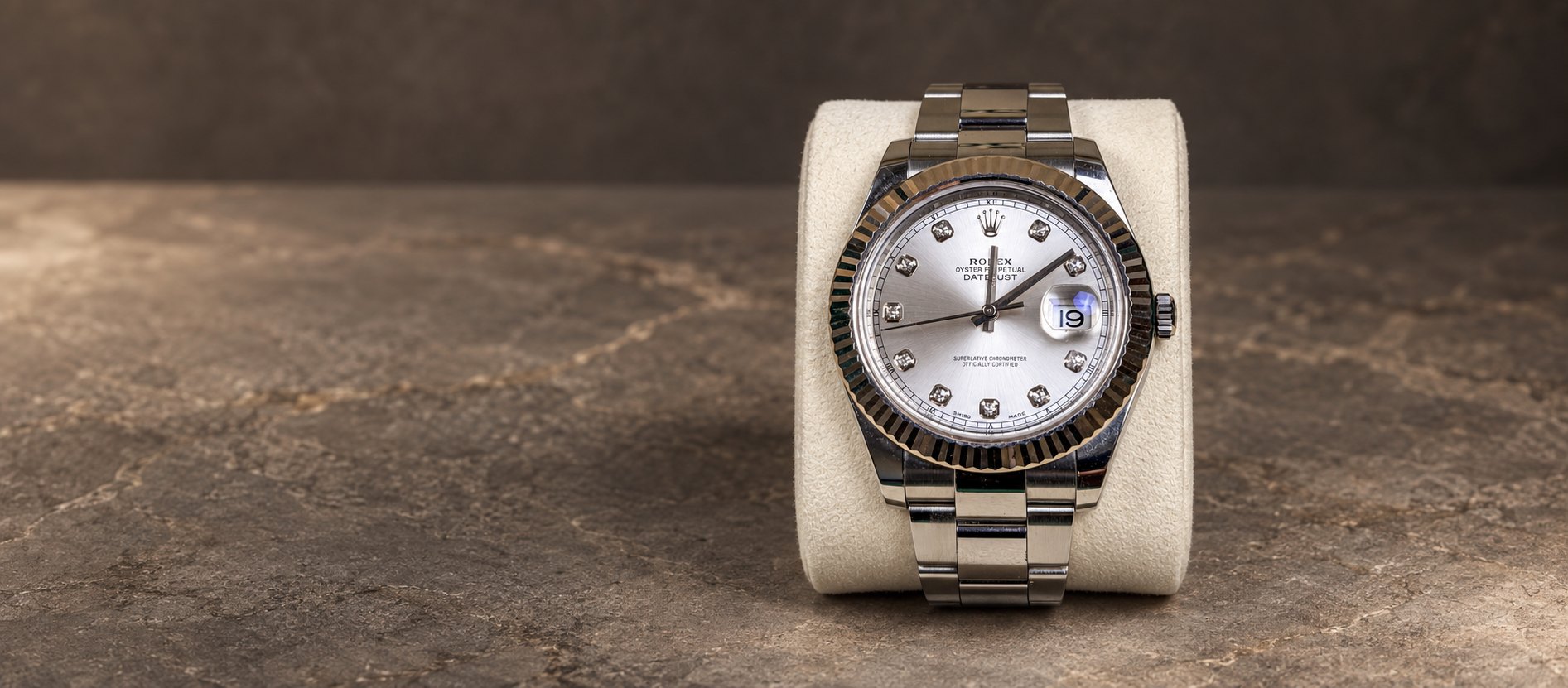 Rolex Datejust II reference 116334 with silver diamond-marker dial, fluted bezel and steel Oyster bracelet on a cream cushion against a warm stone background.