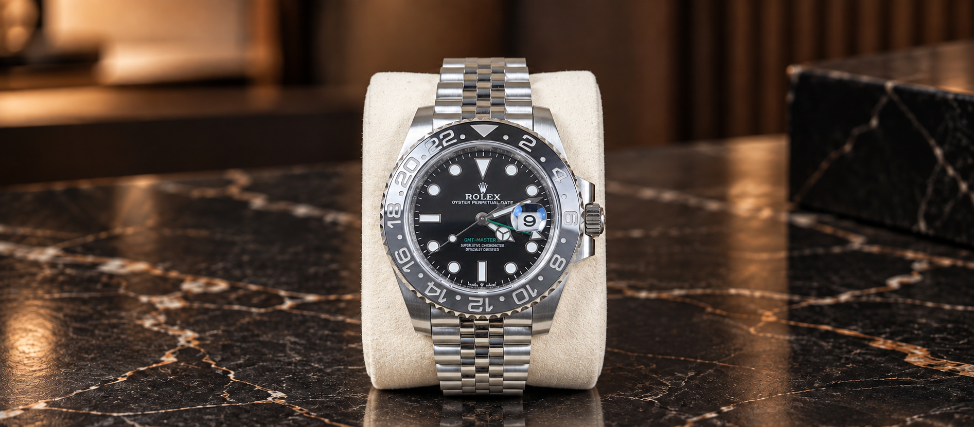 Rolex GMT-Master II 126710GRNR with grey-and-black bezel, black dial and Jubilee bracelet on a cream cushion against a dark stone background.